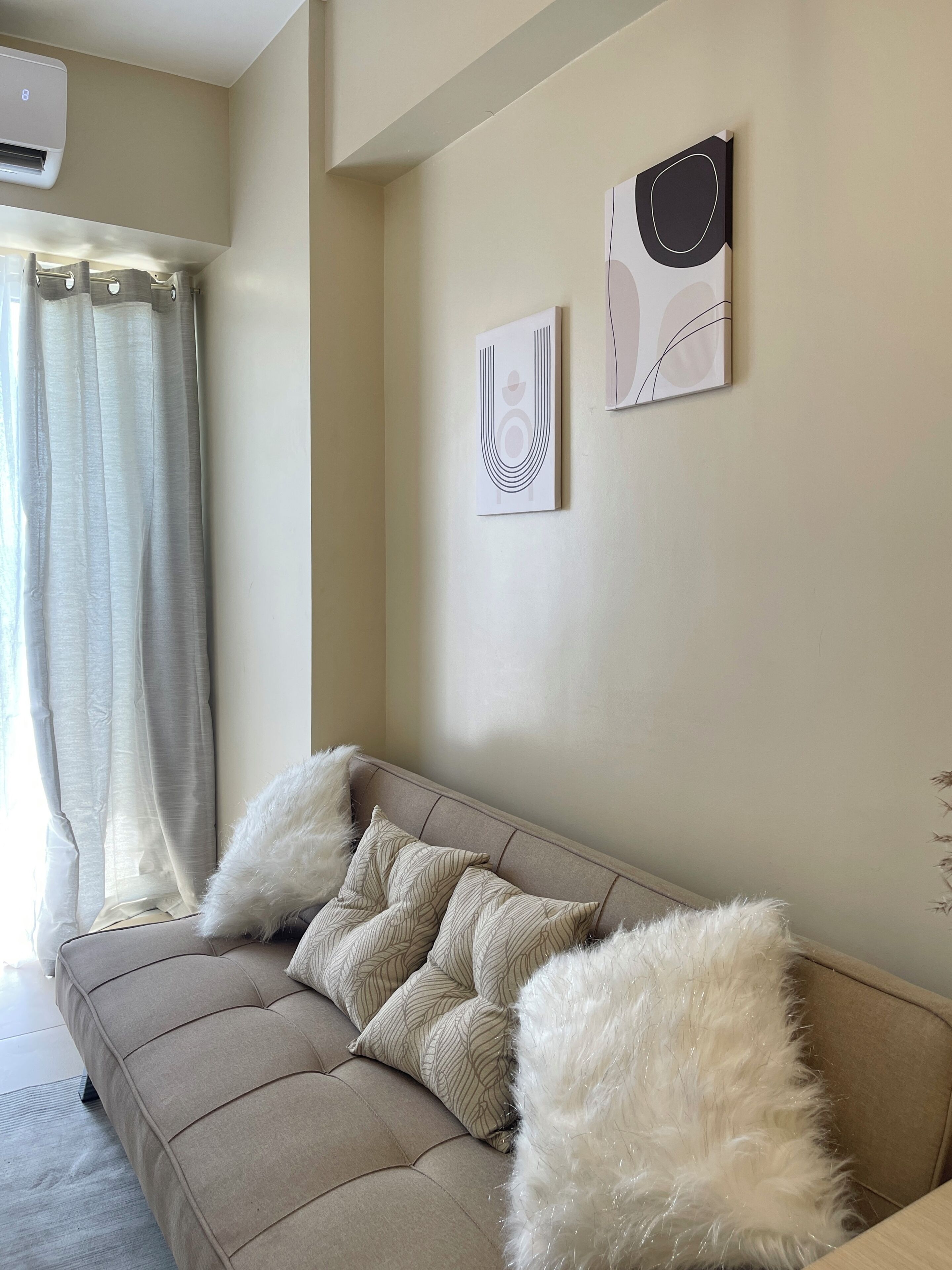 A beige sofa with white and patterned cushions, a wall-mounted air conditioner, and abstract wall art.