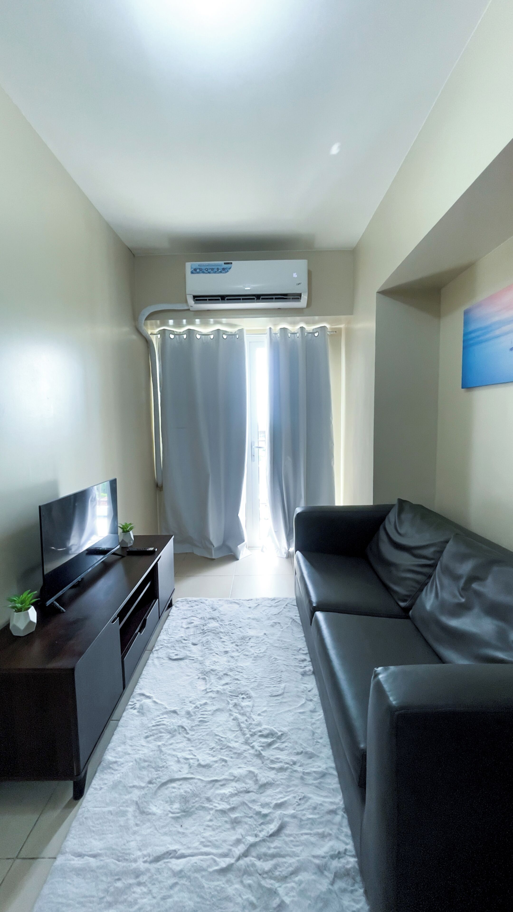 A modern living room with a flat-screen TV, a black leather sofa, a white rug, and a ceiling-mounted air conditioner.