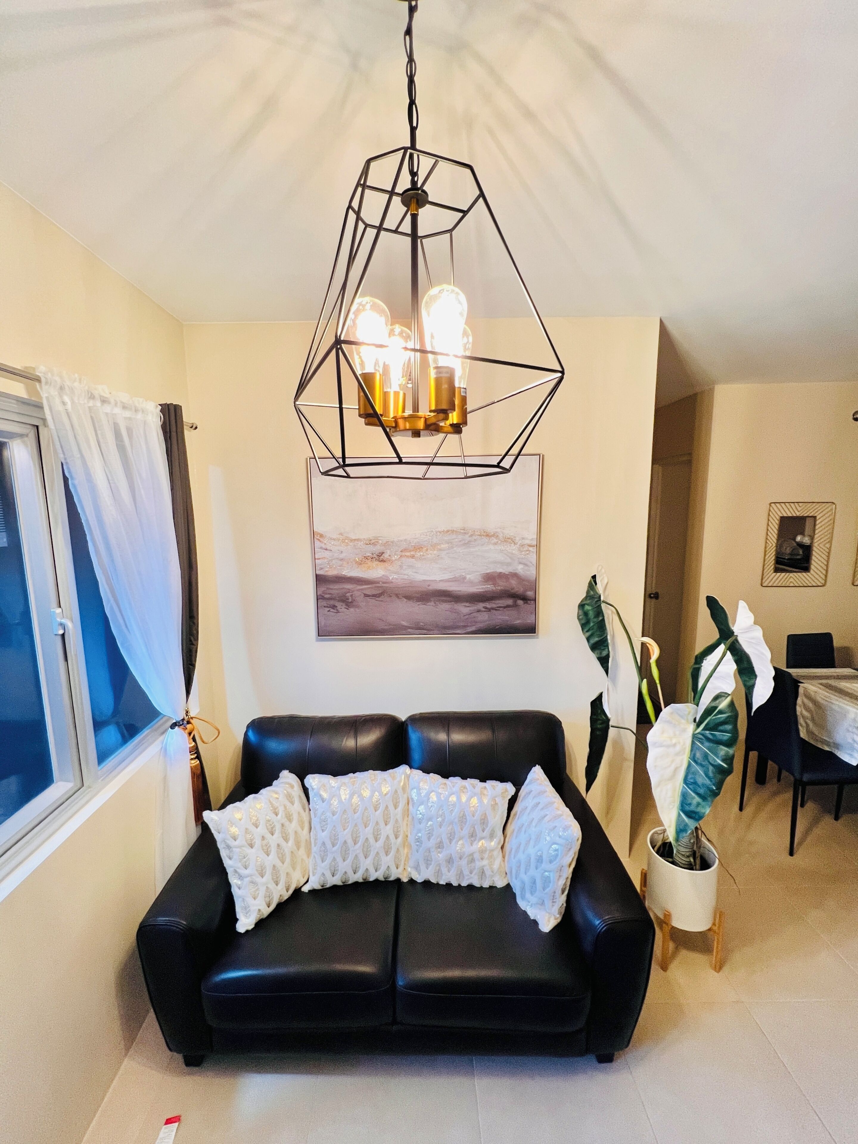 A living room with a black leather sofa, a geometric chandelier, a painting, and a dining area with a table and chairs.