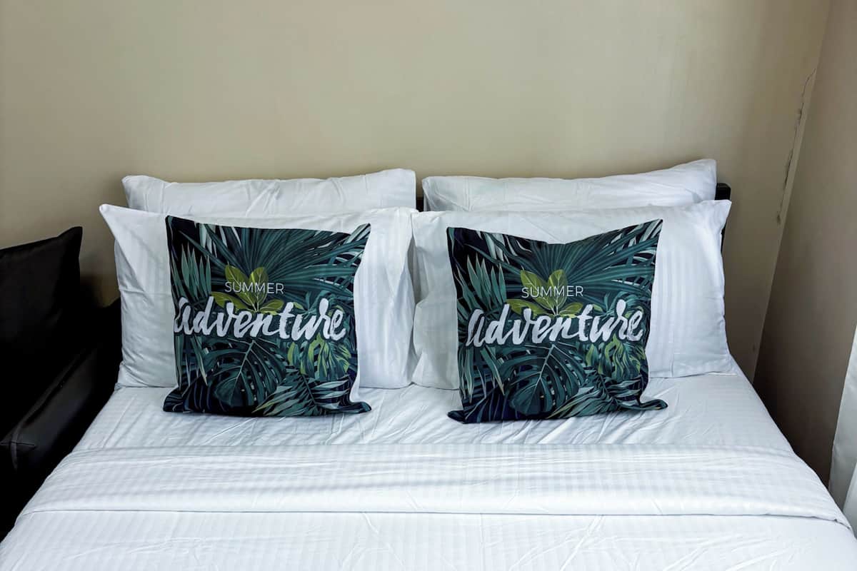 A bed with white bedding and two decorative pillows with tropical leaf patterns and the word 'adventure.'