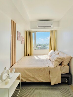 A hotel room with a bed, a white bedside table with a vase and a small sculpture, a window with curtains, and an air conditioning unit. - Avida Towers Atria (Iloilo)