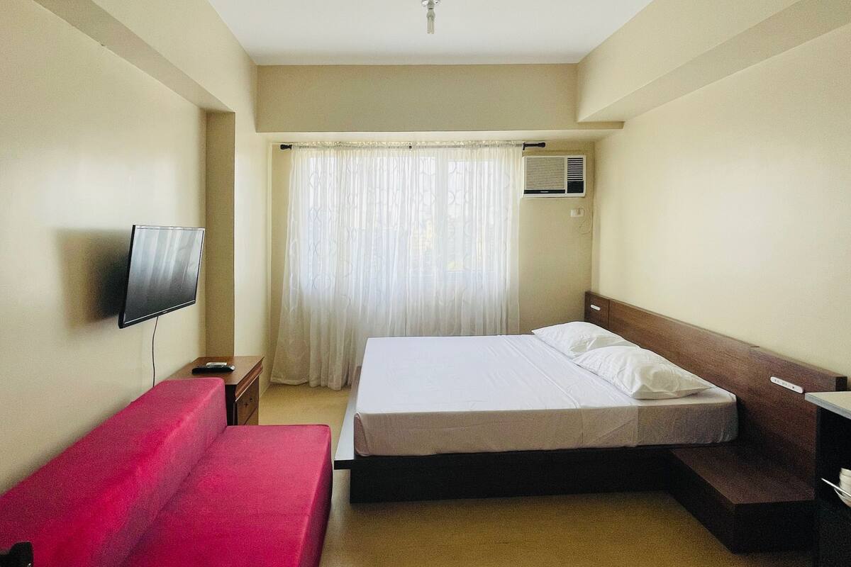 A hotel room with a bed, a TV mounted on the wall, a red sofa, and a window with curtains.