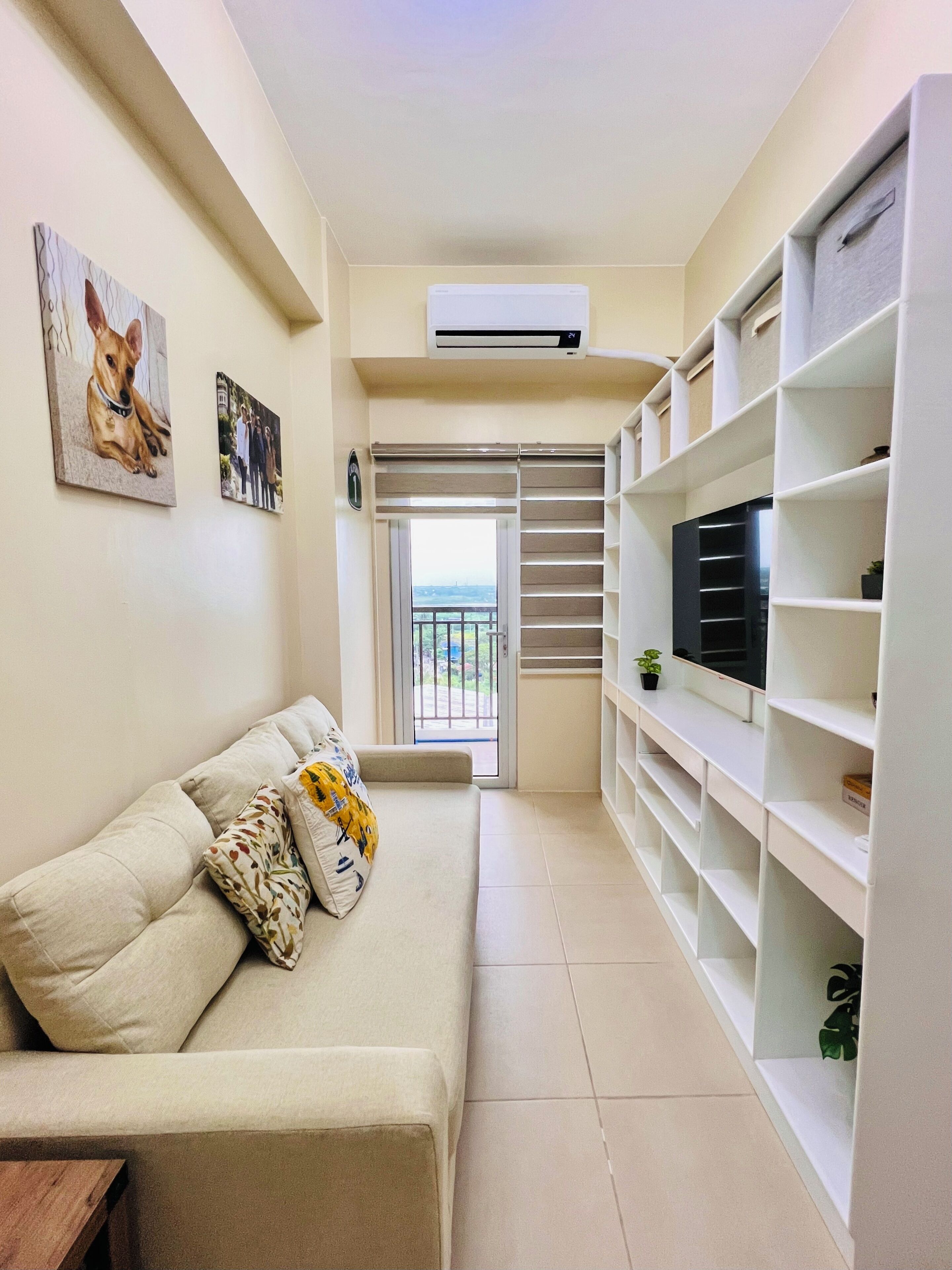 A living room with a beige sofa, a TV on a white shelf, and a balcony door.