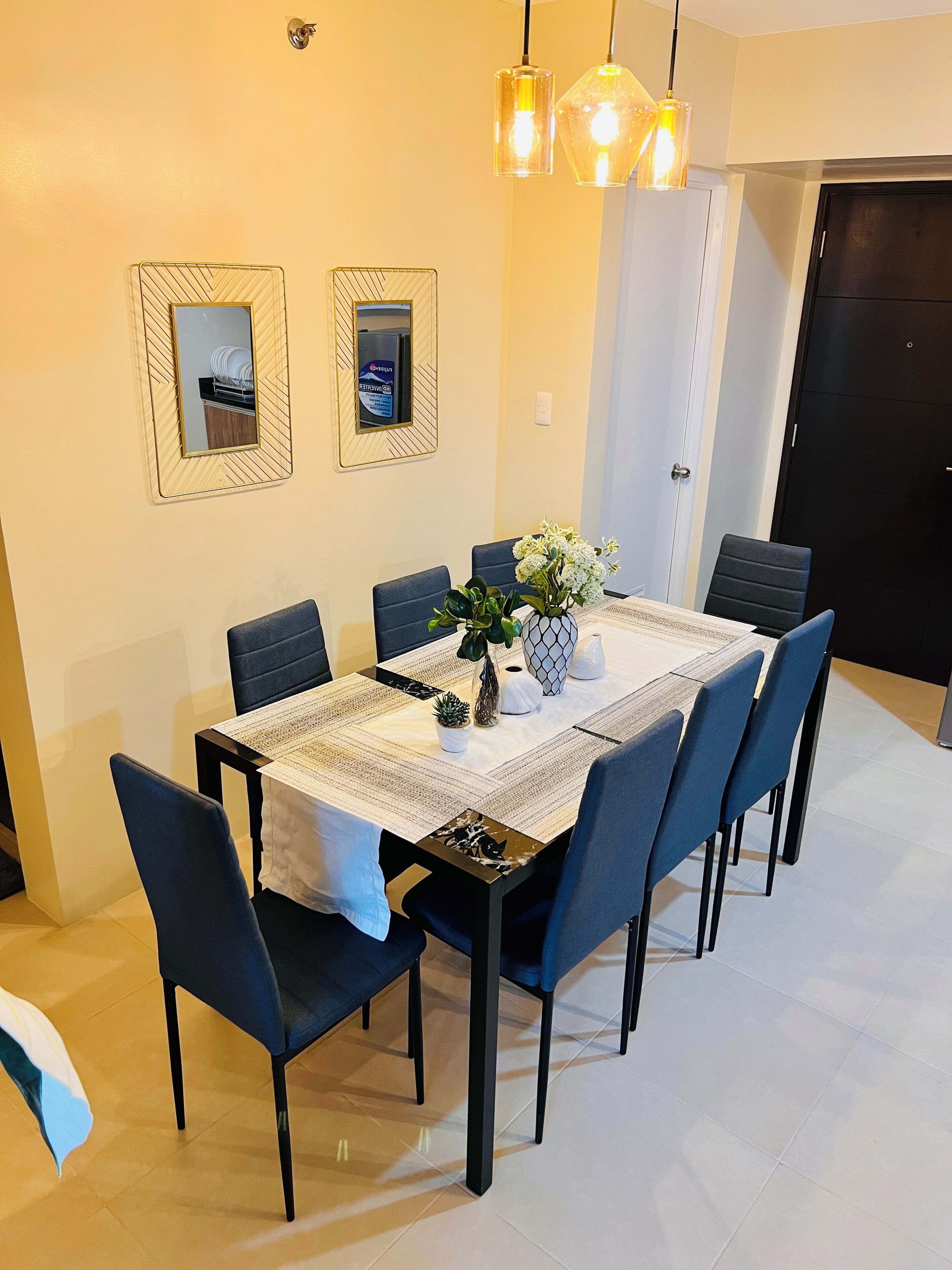 Un comedor con una mesa rectangular, seis sillas y un jarrón con flores. Se observan dos espejos en la pared y una puerta al fondo.
