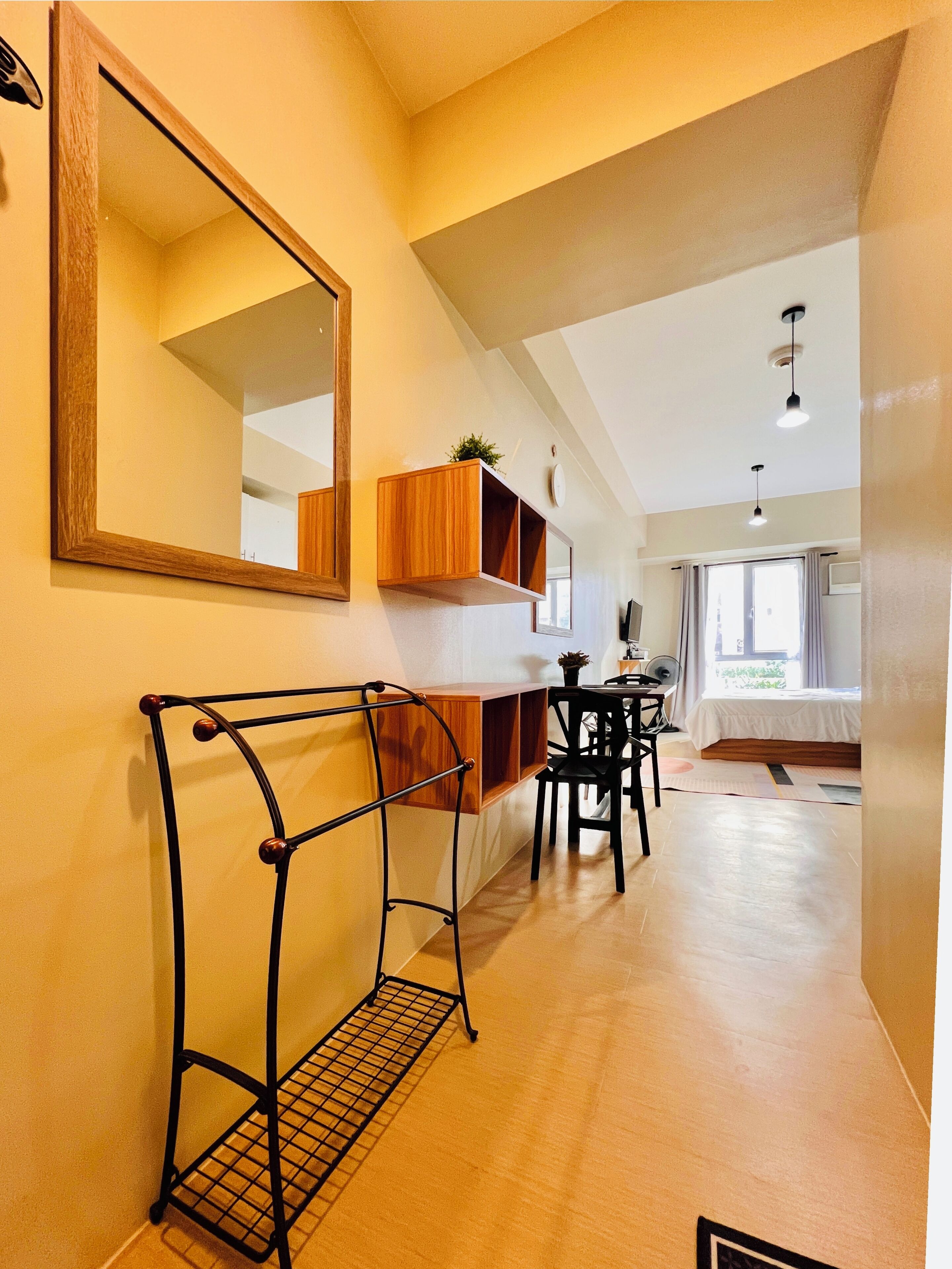 A hallway with a wooden console table, a black metal rack, and a mirror. There is a window with curtains and a bed in the background.