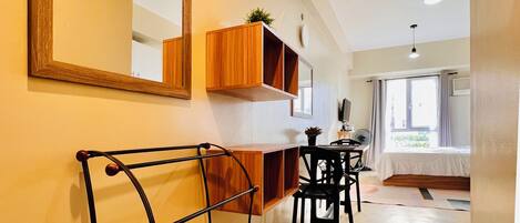 A hallway with a wooden console table, a black metal rack, and a mirror. There is a window with curtains and a bed in the background.