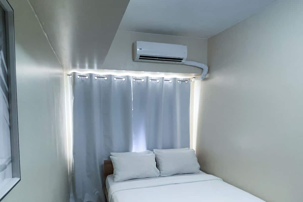 A small, neatly arranged bedroom with a single bed, an air conditioning unit, and a window with curtains.
