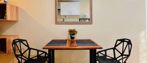 A dining area with a wooden table, two black chairs, a small potted plant, a wall clock, and a mirror reflecting a kitchen area.