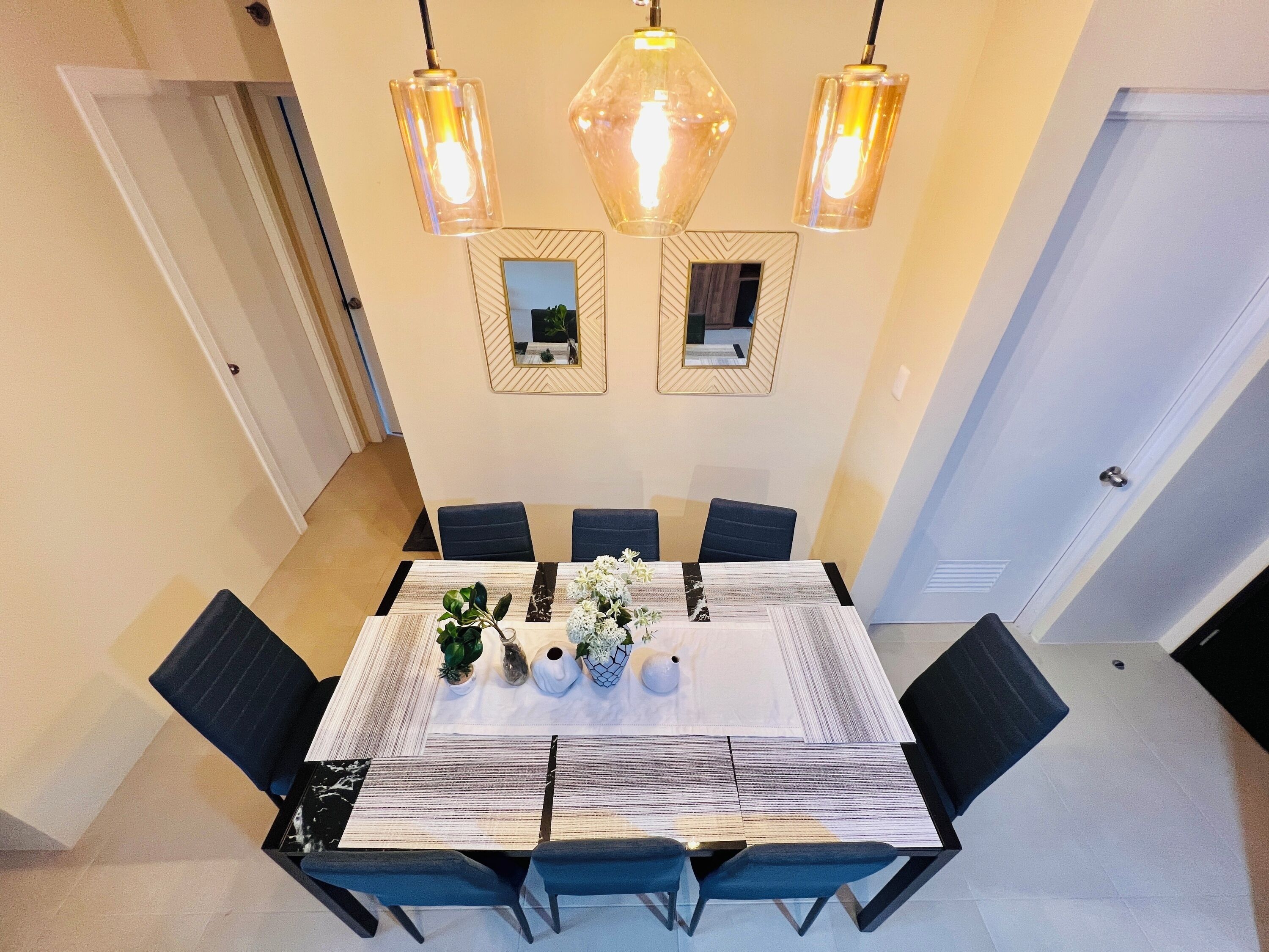 A dining area with a rectangular table, six chairs, and three pendant lights. There are two wall-mounted mirrors and a door in the background.