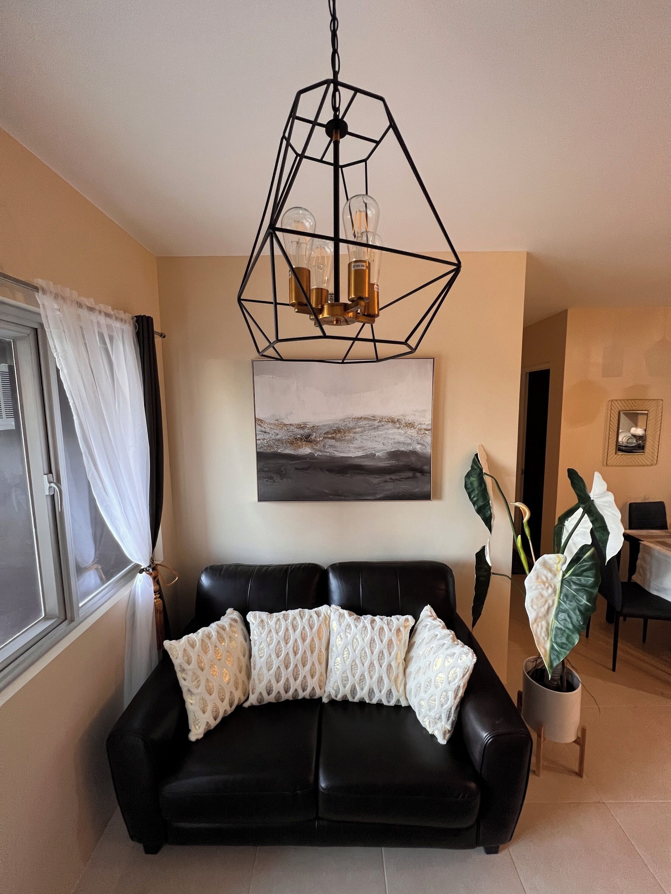 A living room with a black leather sofa, a geometric pendant light, a painting of a landscape, and a potted plant.