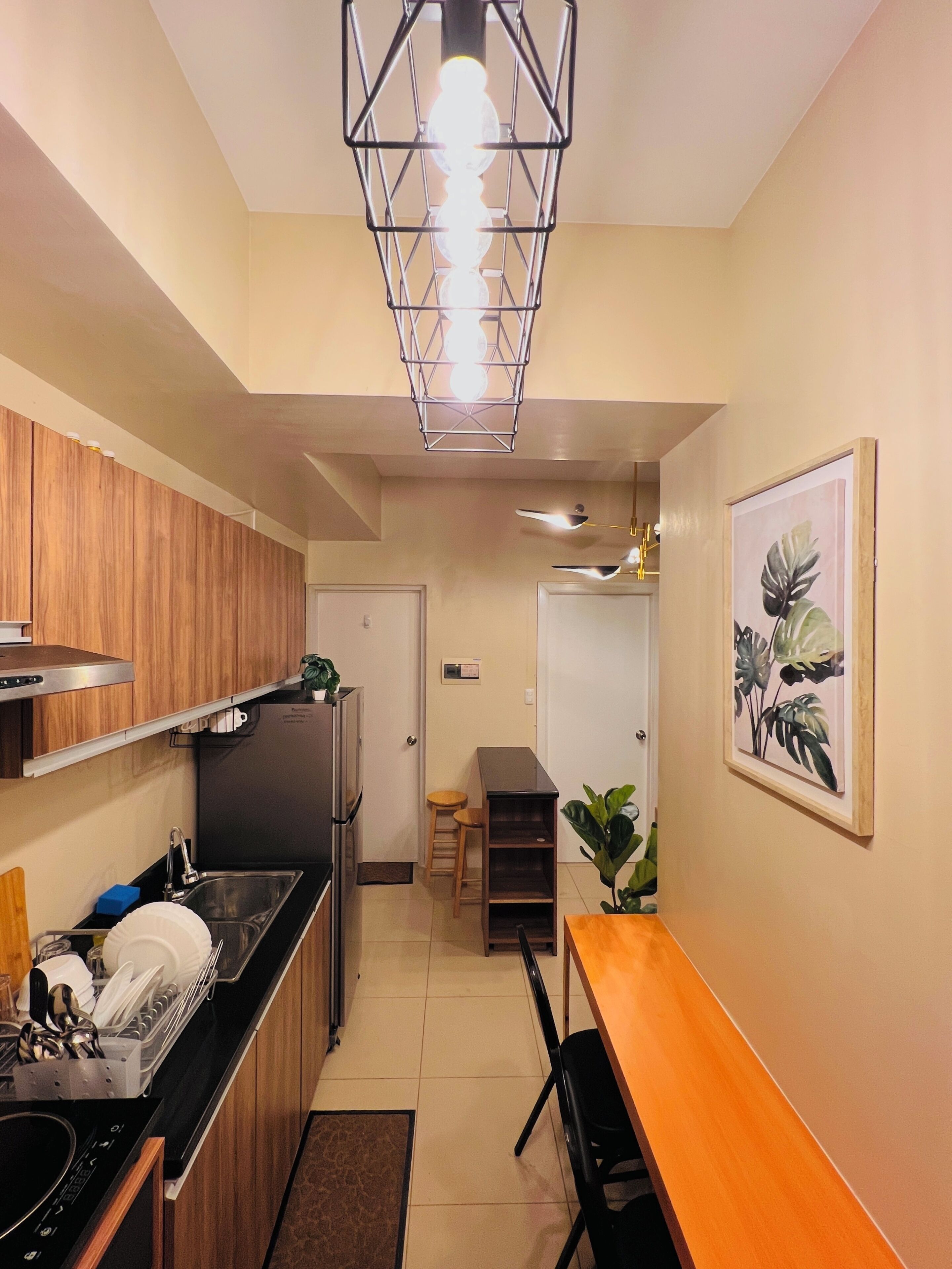 Cocina compacta con armarios de madera, encimera negra y pileta de acero inoxidable. Cuenta con un comedor con mesa y sillas de madera, y una planta en el suelo.