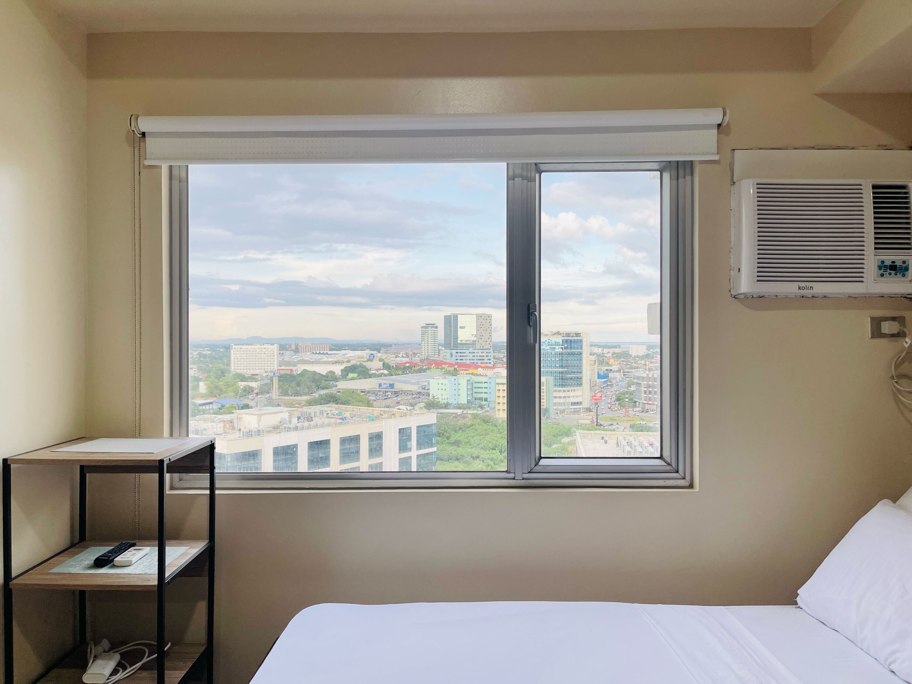 Habitación de hotel con cama, una mesita con un control remoto y una ventana con vista a la ciudad.