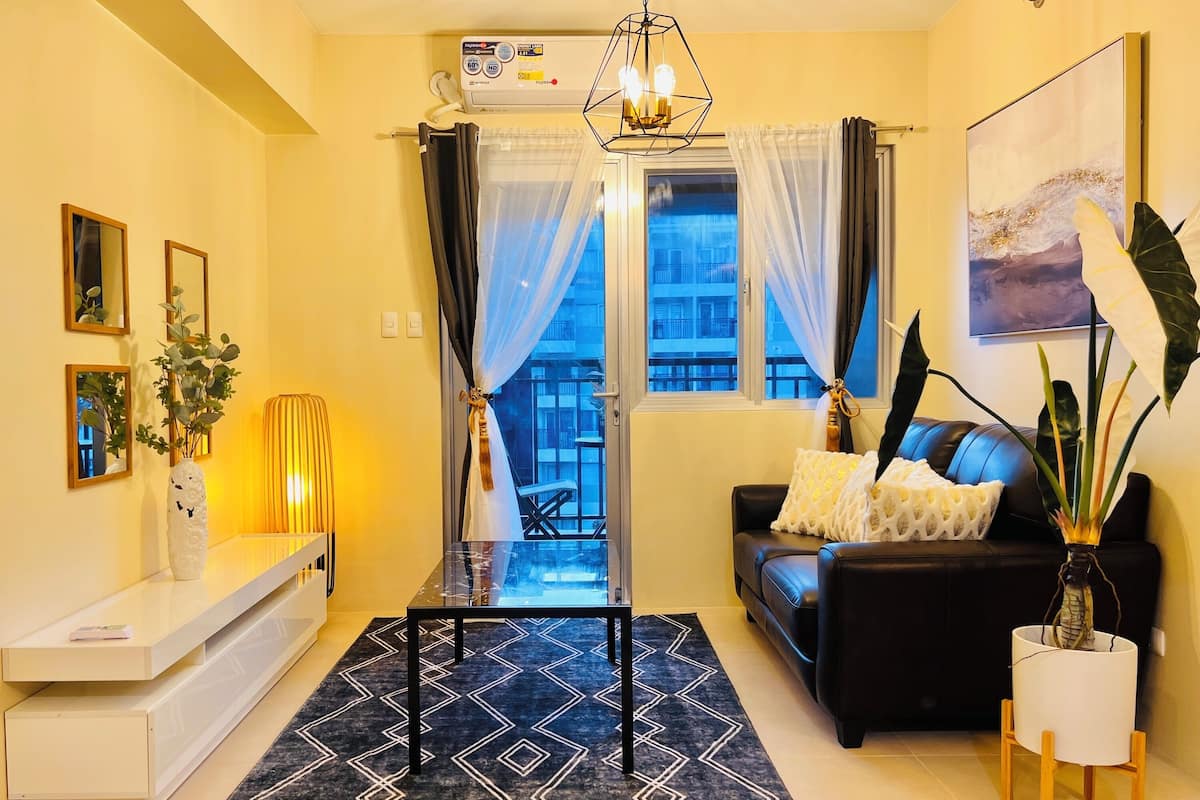 A modern living room with a black sofa, a coffee table, a white console table, and a large window with curtains.