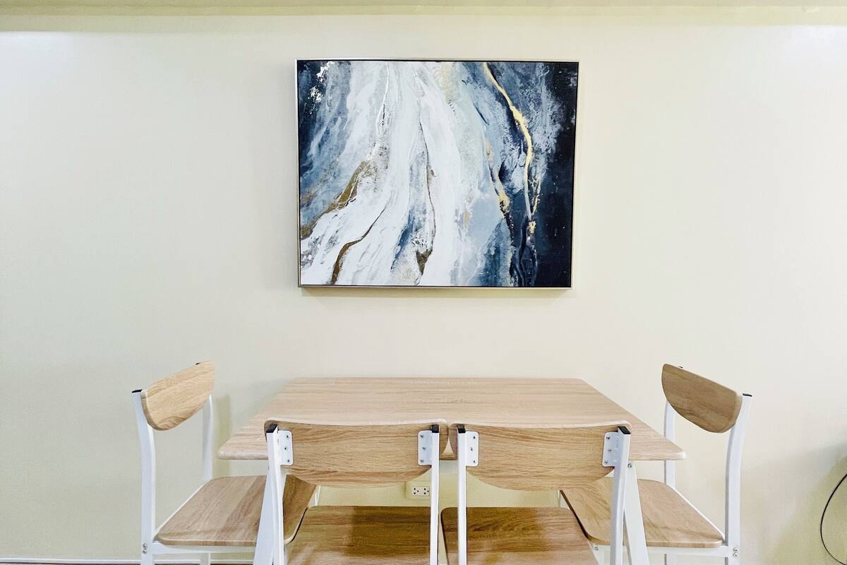 A dining area with a wooden table and white chairs, a painting on the wall, and a light-colored floor.