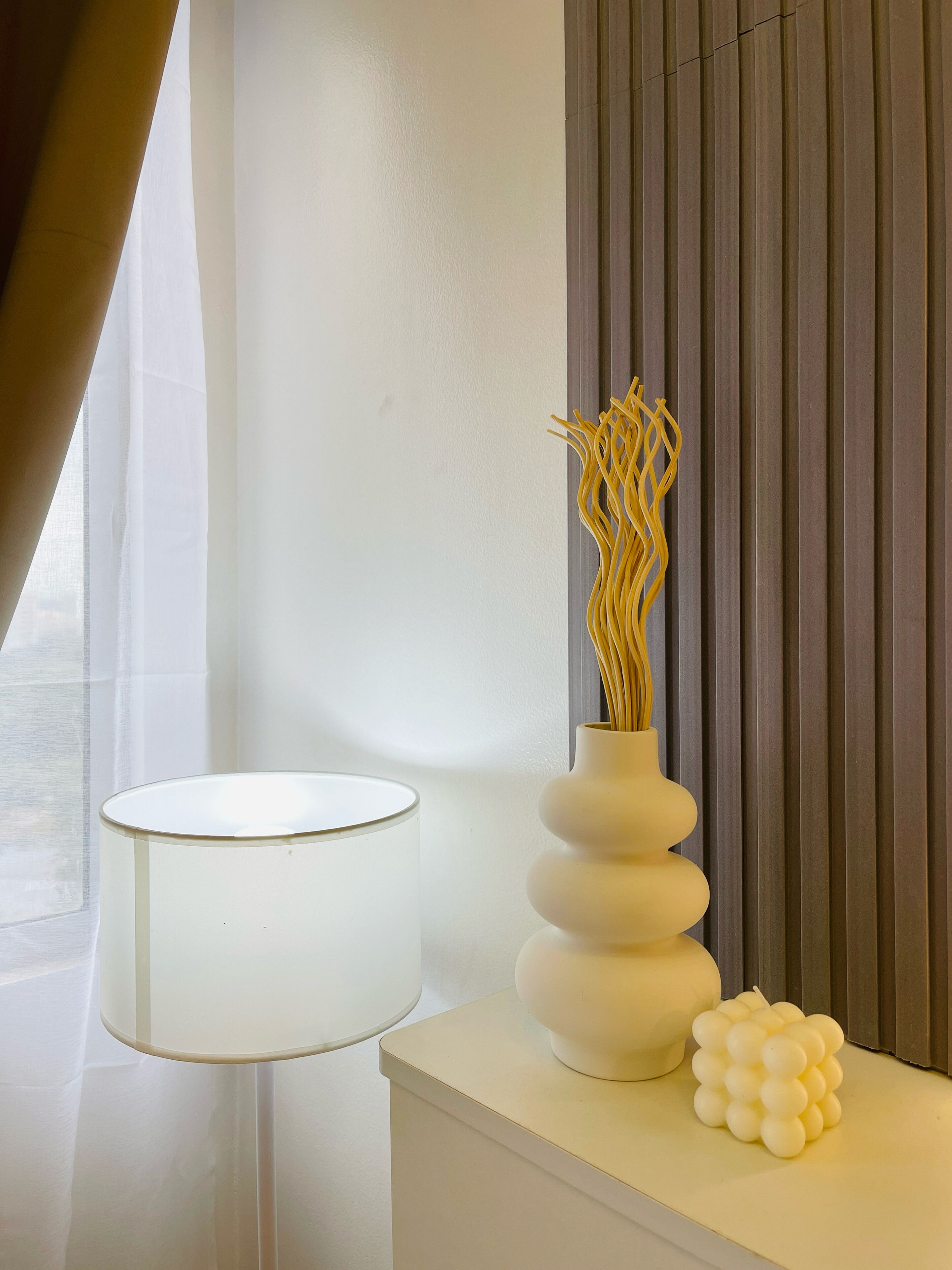 A modern room with a white lamp, a vase with dried plants, and a decorative white structure on a white console table.