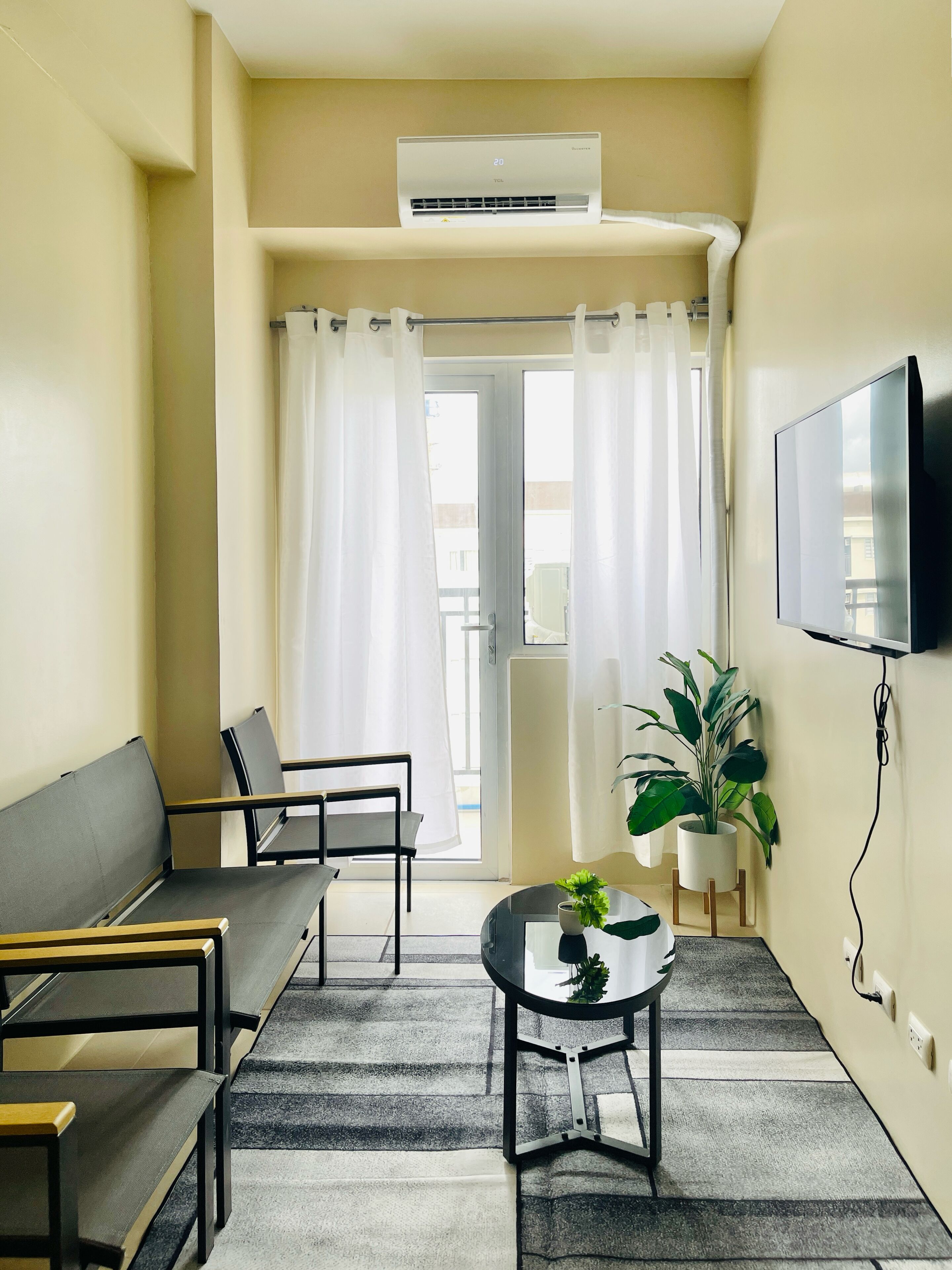 A small, modern room with a sofa, a table, a TV mounted on the wall, and an air conditioning unit above the door.