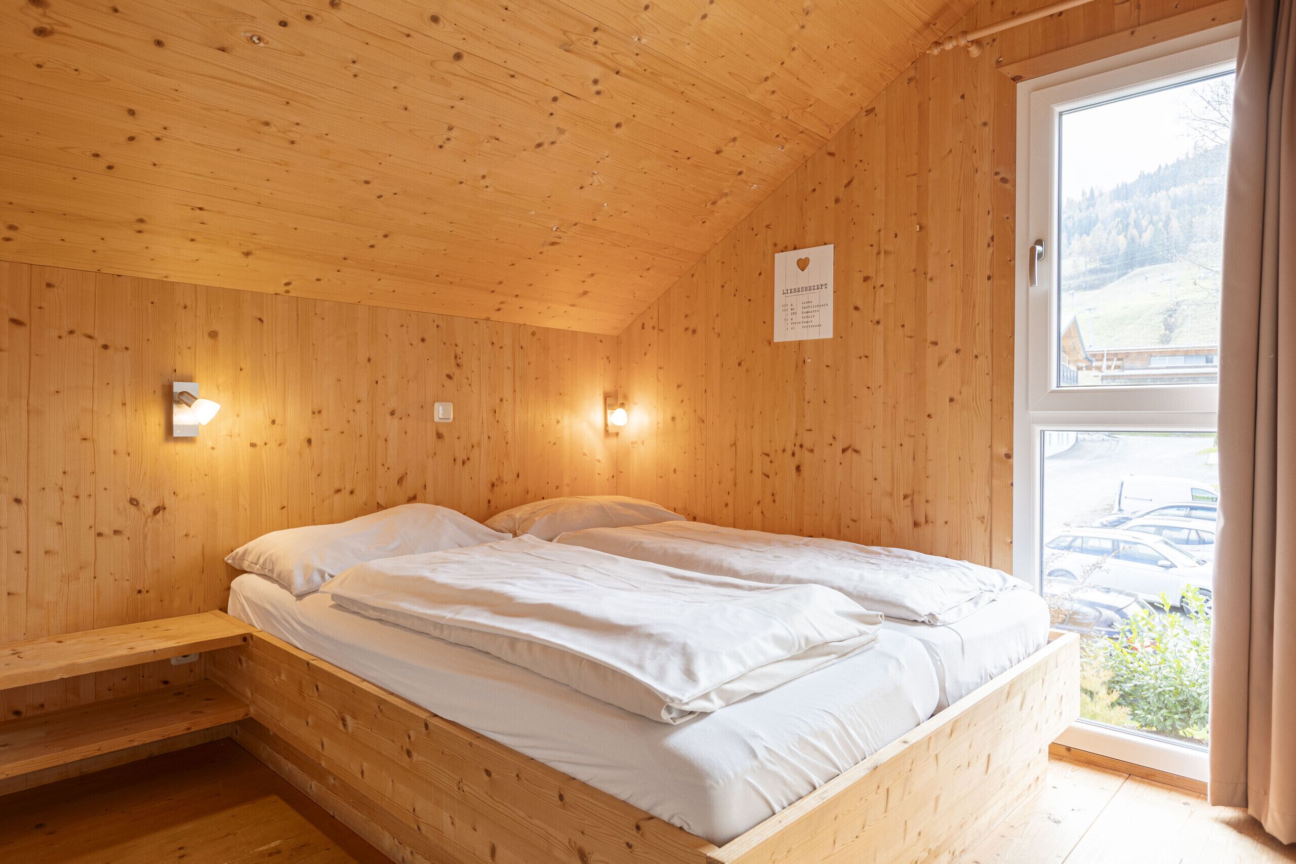 A wooden cabin room with a bed, a small table, and a window with a view of a parking area.