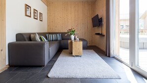 Una sala moderna con un sofá seccional gris, una mesa de centro de madera con un jarrón de flores, una alfombra gris y un ventanal con cortinas.