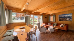 A wooden interior with a dining table, chairs, a sofa, and a large window.