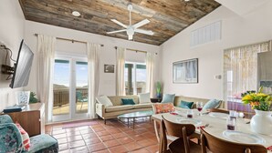 A spacious living area with a wooden ceiling, a dining table set for four, and a balcony with a view. - Seaside Cottages, 3 Homes, Pool, Beach & Golf Cart (Port Aransas)