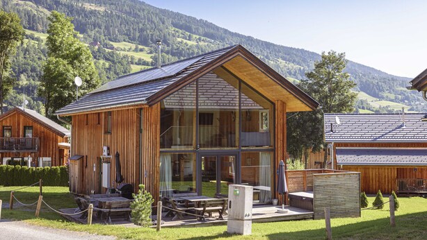 A wooden lodge with large windows, a patio with tables and chairs, and a view of the surrounding mountains.