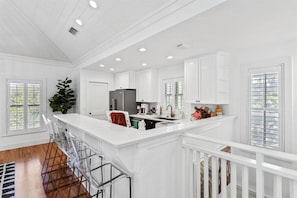A spacious kitchen with white cabinetry, a central island with bar stools, and a large window providing natural light. - Beach Cottage | Seacrest | Private Beach Access (Seacrest)