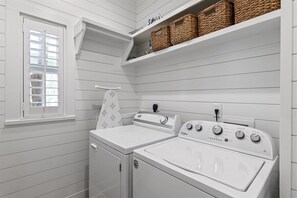 A laundry room with a front-loading washer and dryer, a window with blinds, and wicker baskets on a shelf. - Beach Cottage | Seacrest | Private Beach Access (Seacrest)