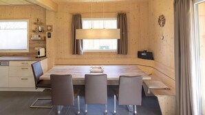 Un interior de madera con una mesa de comedor, cuatro sillas, una ventana con cortinas, un reloj y un estante con objetos.