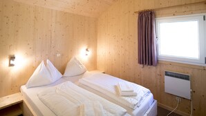 Puinen huone, jossa on sänky, tyynyjä, ikkuna verhoilla ja seinään kiinnitetty lämmitin.