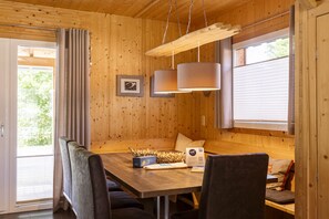 Un intérieur en bois comprenant une table à manger, des chaises et un banc. On y trouve deux lampes suspendues, une fenêtre avec des stores et une porte donnant sur l’extérieur.