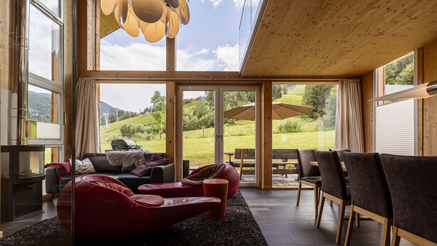 Ett modernt vardagsrum med en röd fåtölj, en soffa och en matplats med stolar. Stora fönster ger utsikt över ett grönskande landskap.