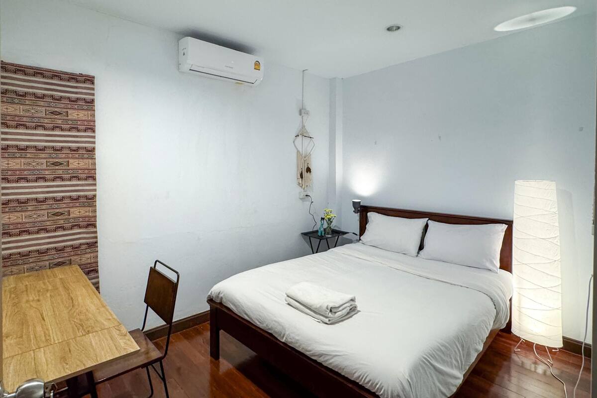 Una habitación de hotel con cama, escritorio, silla, aire acondicionado de pared, lámpara de noche y un adorno en la pared.