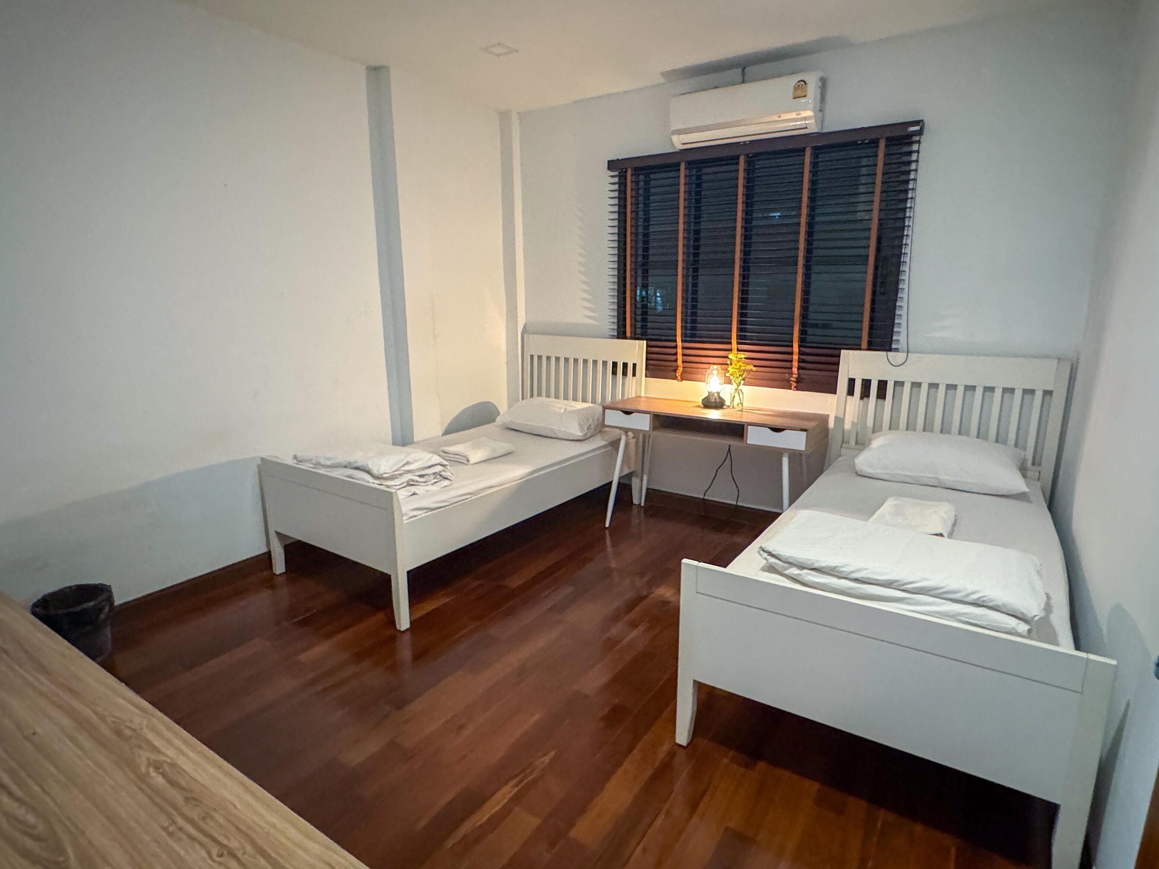 A small, clean room with two single beds, a desk, and a window with blinds.