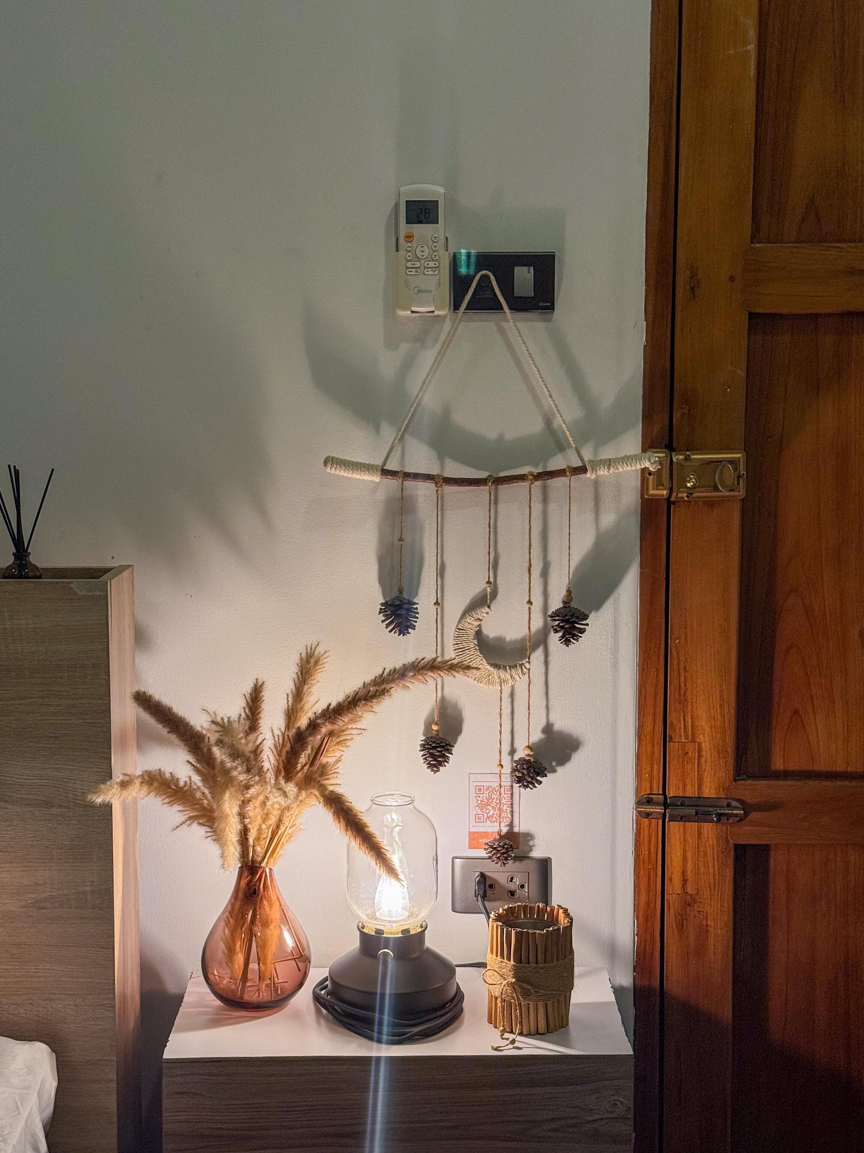 Ein Nachttisch mit Lampe, einer Vase mit Trockenblumen, einem kleinen Dekorationsgegenstand und einem an der Wand montierten Gerät.