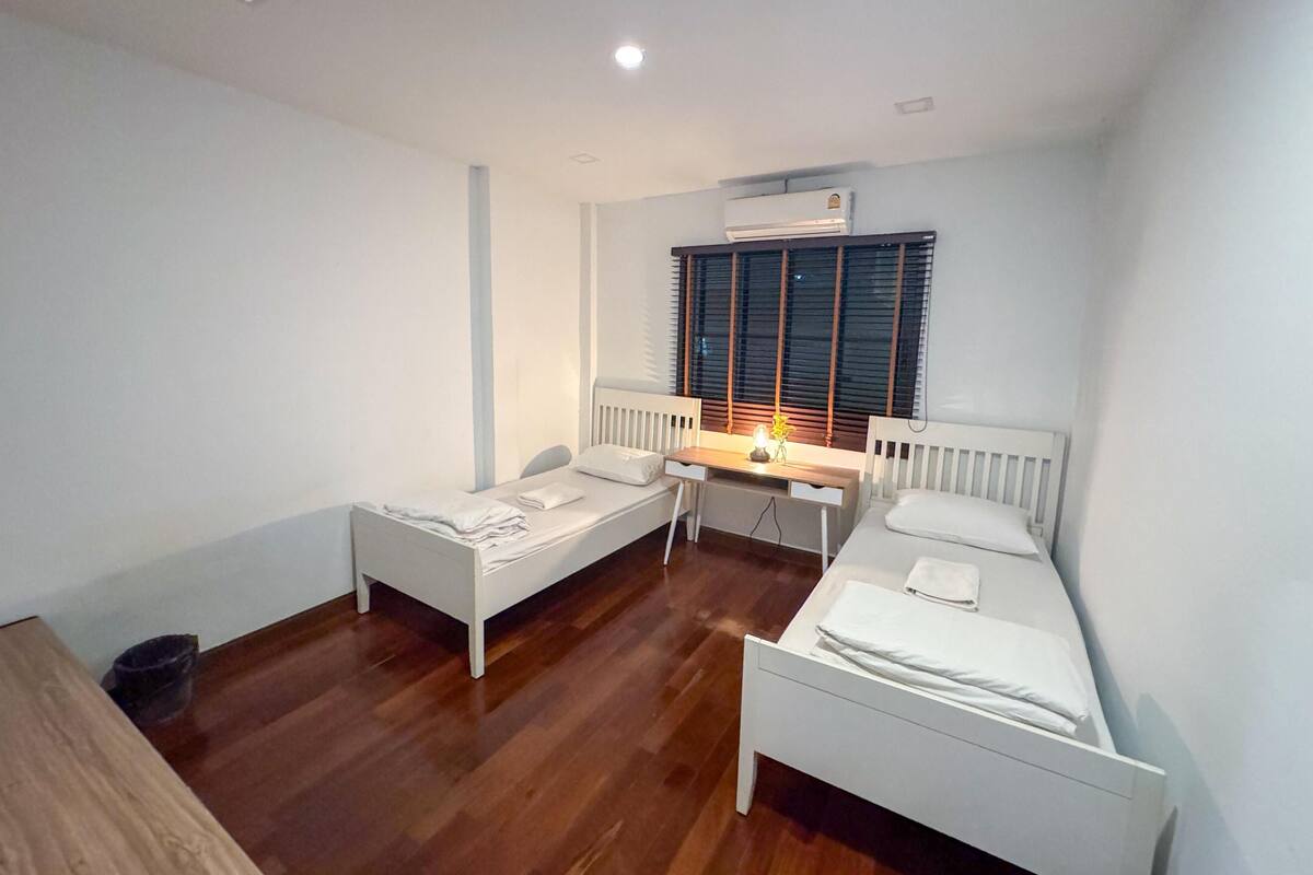 Una habitación pequeña y limpia con dos camas individuales, un escritorio y una ventana con persianas.