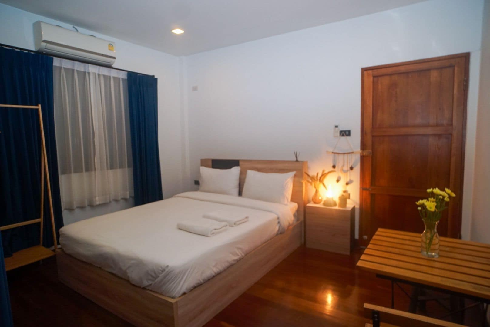 A hotel room with a wooden bed, a nightstand with a lamp, a wooden table, and a wooden wardrobe.