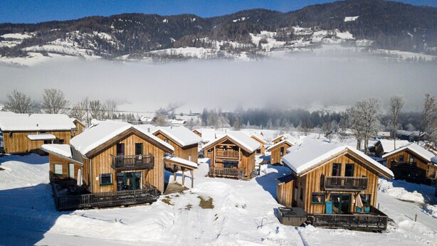 Una hilera de cabañas de madera con balcones, rodeadas de nieve y montañas al fondo.