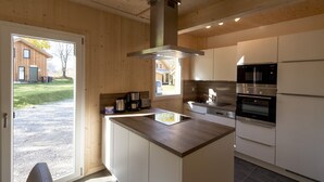 A modern kitchen with a central island, stainless steel appliances, and a view of a wooden building outside.