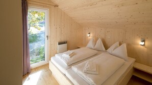 A wooden cabin bedroom with a large bed, two bedside tables, a window with curtains, and a heater.