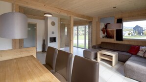 A spacious living area with a wooden ceiling, a sofa set, a dining table with chairs, and a large window offering a view of the outdoors.