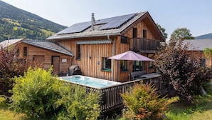 A wooden cabin with a hot tub and a balcony, surrounded by greenery and mountains.