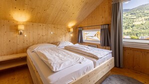 A wooden cabin bedroom with two beds, a window with curtains, and a view of a forested landscape.