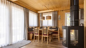 A cozy cabin interior with a wooden dining table, chairs, a fireplace, and large windows with curtains.