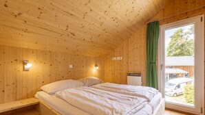 Une pièce de cabane en bois comprenant un lit, une fenêtre donnant sur l’extérieur et permettant d’apercevoir un véhicule, et un luminaire fixé au mur.