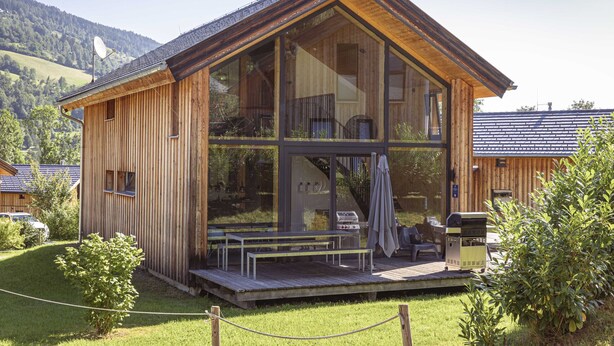 A wooden cabin with large windows, a deck, and a barbecue grill.
