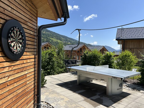 Une cible de fléchettes fixée à un mur en bois, une table de ping-pong et, en arrière-plan, une vue sur des maisons en bois et des montagnes.