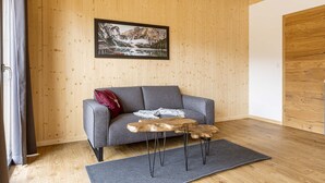 A modern living room with a wooden wall, a grey sofa, a wooden coffee table, and a framed photograph of a mountain landscape.