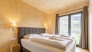 A bedroom with a wooden ceiling, a bed with white linens, two wall-mounted lamps, and a view of a green landscape through a glass door.
