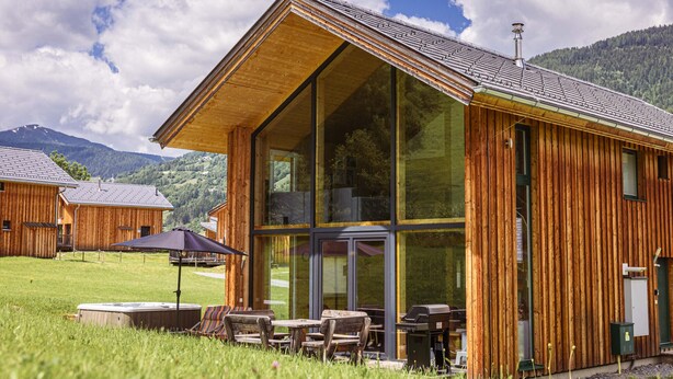 A wooden cabin with large windows, a patio with outdoor furniture, and a mountainous backdrop.