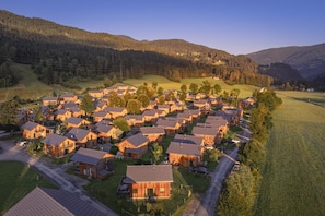A residential area with multiple wooden houses, a central road, and surrounding green fields and hills.